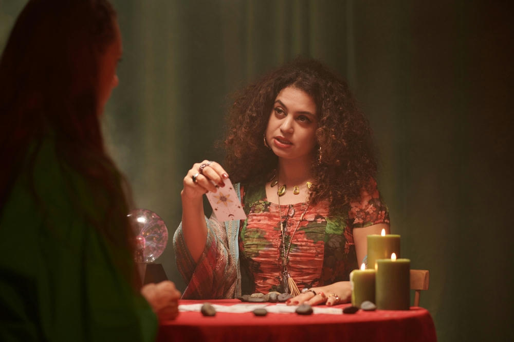A tarot cards reader talking to a client. Both are sitting at a table with candles and a crystal ball.