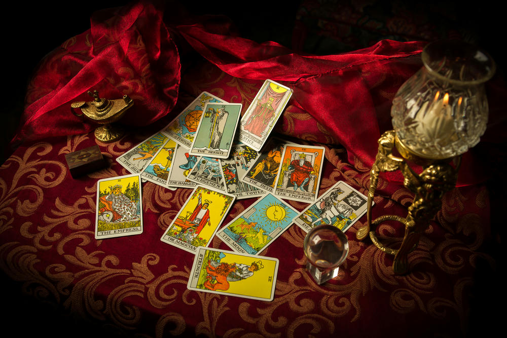 A pile of tarot cards lies scattered and spread across a tabletop surrounded by multiple occult items.