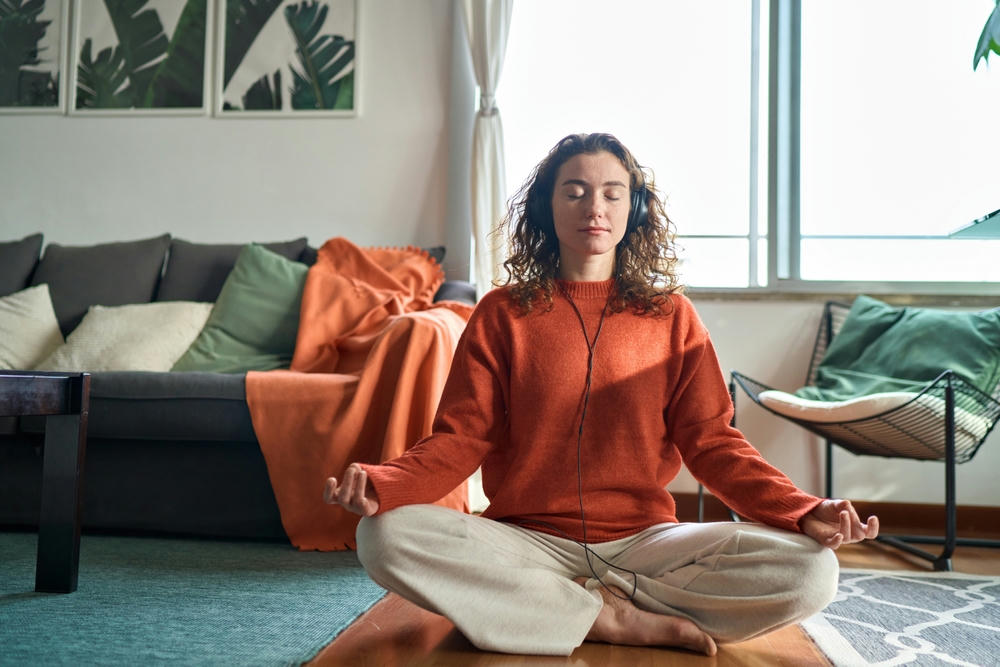 A healthy mindful woman wearing headphones listening to calming audio sitting on the floor at home doing meditation.