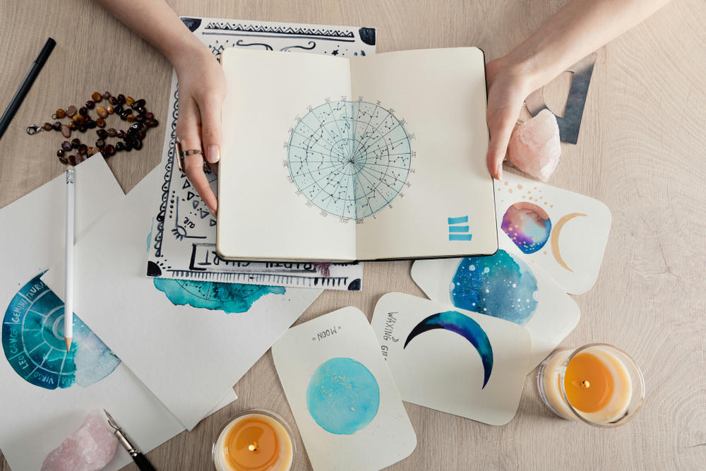 Top view of astrologer holding a notebook with watercolor drawings and zodiac signs on cards on the table.