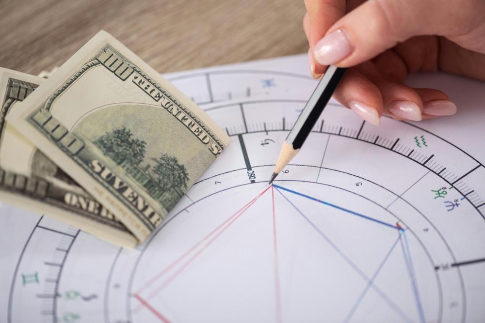 Cropped view of astrologer drawing natal chart beside dollar banknotes on the table.