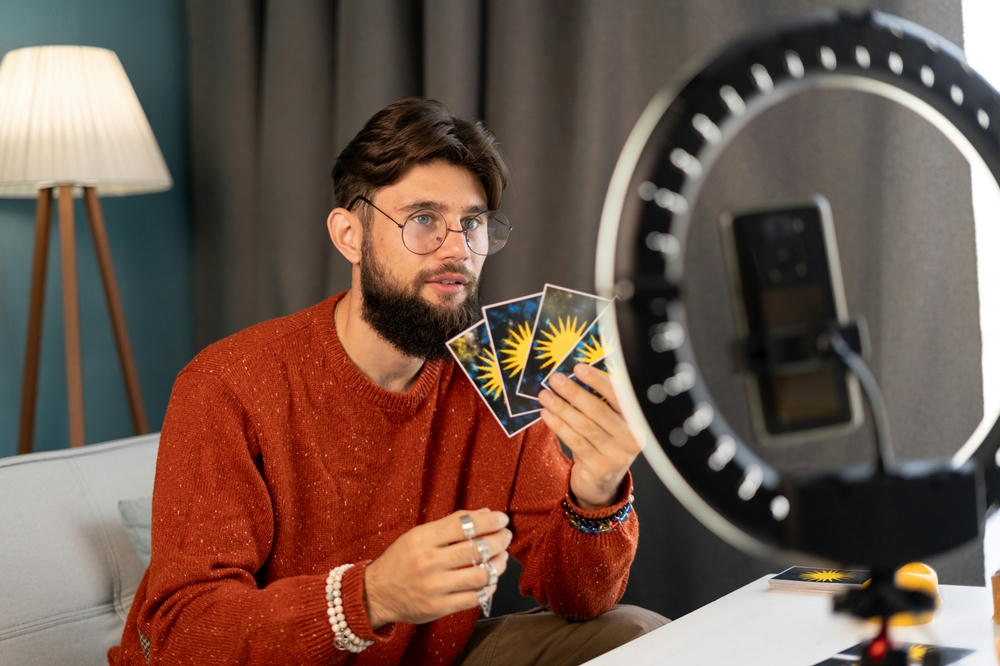 Young male fortune-teller with tarot cards recording video for blog at home.