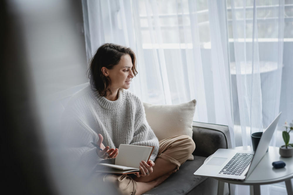  Female student in a sweater studies at home online sitting on the couch making notes in a notebook.
