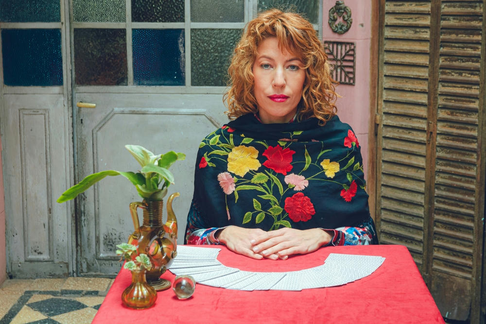 Elegant tarot reader with blonde hair, sitting looking at the camera with a deck of cards fanned out on the table